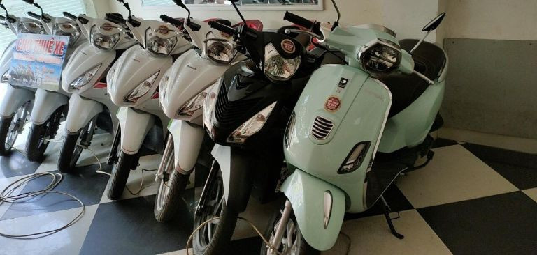Một người đàn ông đang xem điện thoại khi ngồi trên xe máy đã thuê, phía sau là khách sạn